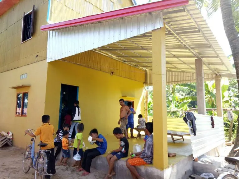 Children sitting on the covered patio beside a yellow house at Kampong Dey Dos